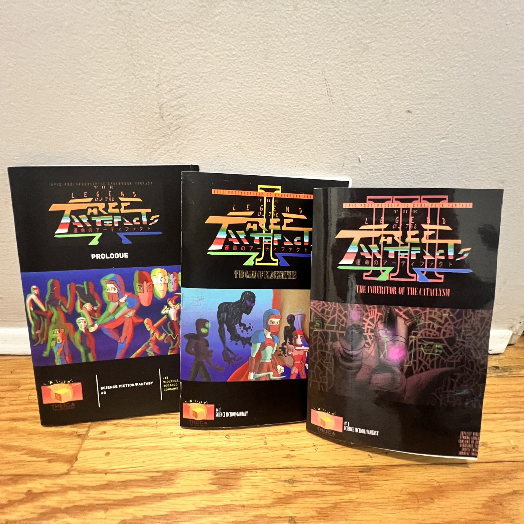 Three issues of The Legend of the Three Artifacts comic displayed upright on a wooden floor, including the prologue and two numbered issues.