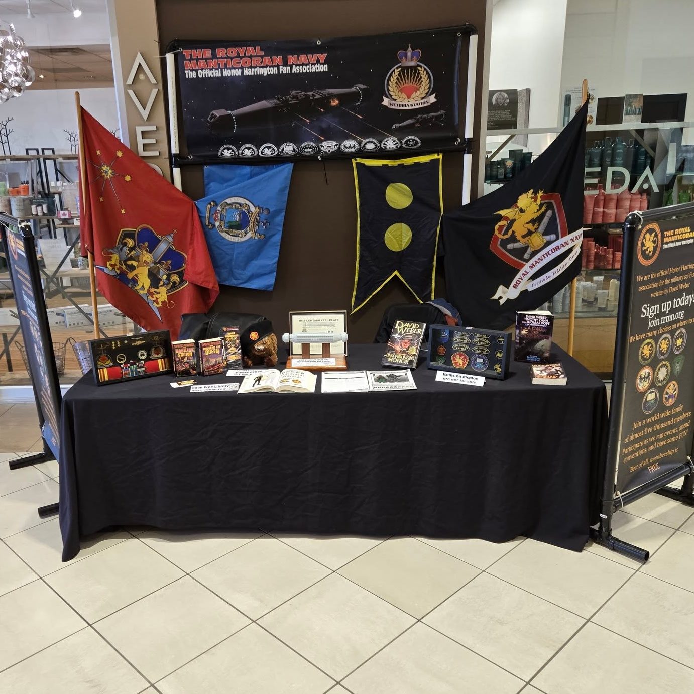 Royal Manticoran Navy fan association display table with Honor Harrington banners, flags, books, props, and informational signs set up in a shopping mall.
