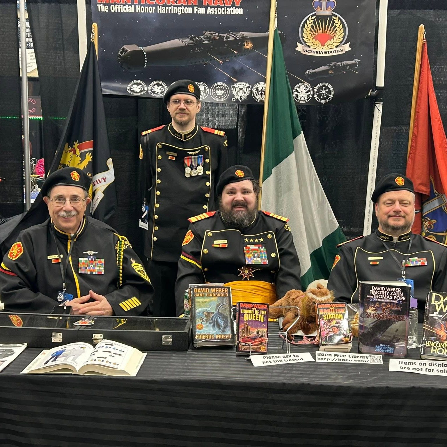 Four Royal Manticoran Navy members in black-and-gold science fiction military-style uniforms seated behind a display table with Honor Harrington books, props, and fan association materials.
