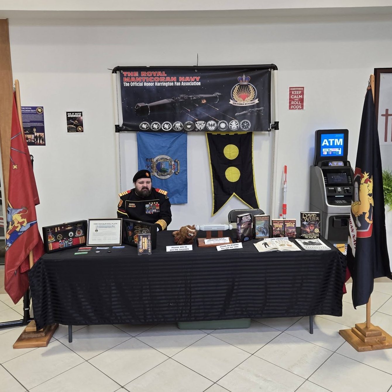 Royal Manticoran Navy display table with flags, banners, Honor Harrington books, props, and a costumed fan association member seated behind the table.