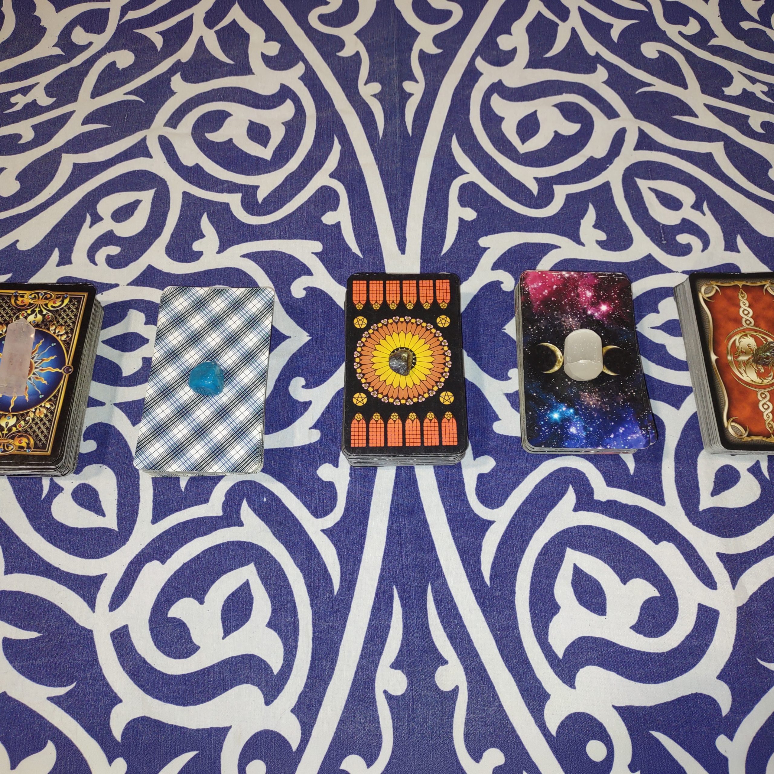 Five tarot card decks arranged on a blue-and-white patterned cloth, each topped with a small crystal or stone.