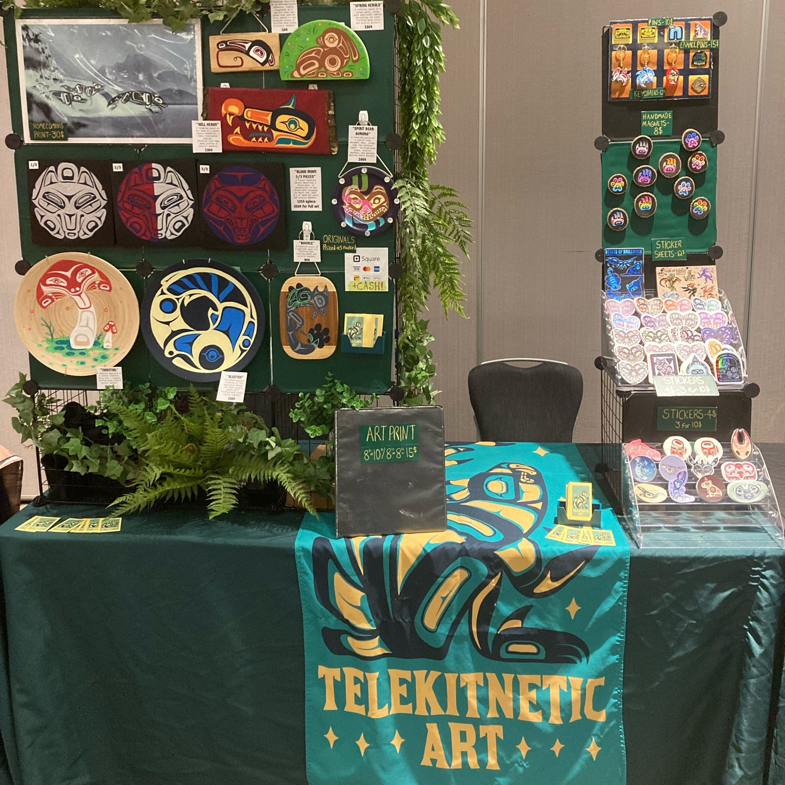 TeleKitnetic Art vendor table display with art prints, stickers, handmade magnets, pins, keychains, painted pieces, greenery, and a teal table banner.