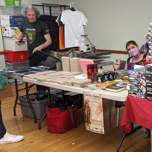 West Coast Speculative Fiction Association table at Fandom Bazaar with books, posters, T-shirts, boxes, and volunteers set up inside a community hall.