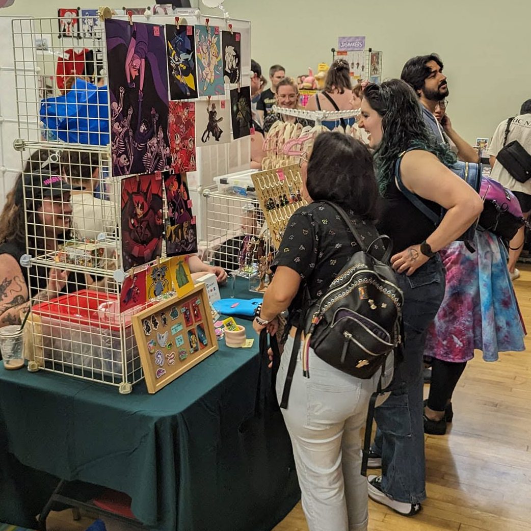 Busy Fandom Bazaar vendor hall with attendees browsing artist tables displaying prints, charms, stickers, pins, and handmade goods.