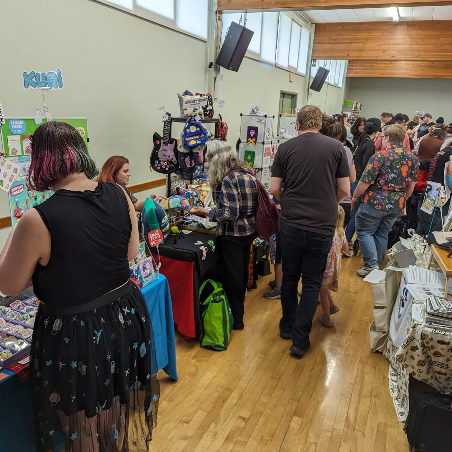 Crowded Fandom Bazaar vendor aisle with attendees shopping at colourful artist and maker tables inside a community hall.