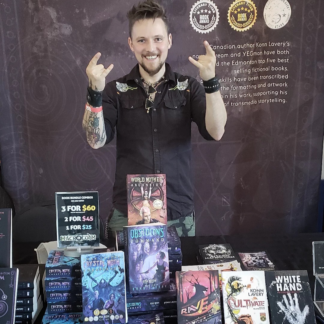 Horror and dark fantasy author Konn Lavery smiling behind a vendor table displaying his books, including Crystal Moth Conspiracy, World Mother Ascension, Obsidian’s Command, Cultivate, and White Hand.