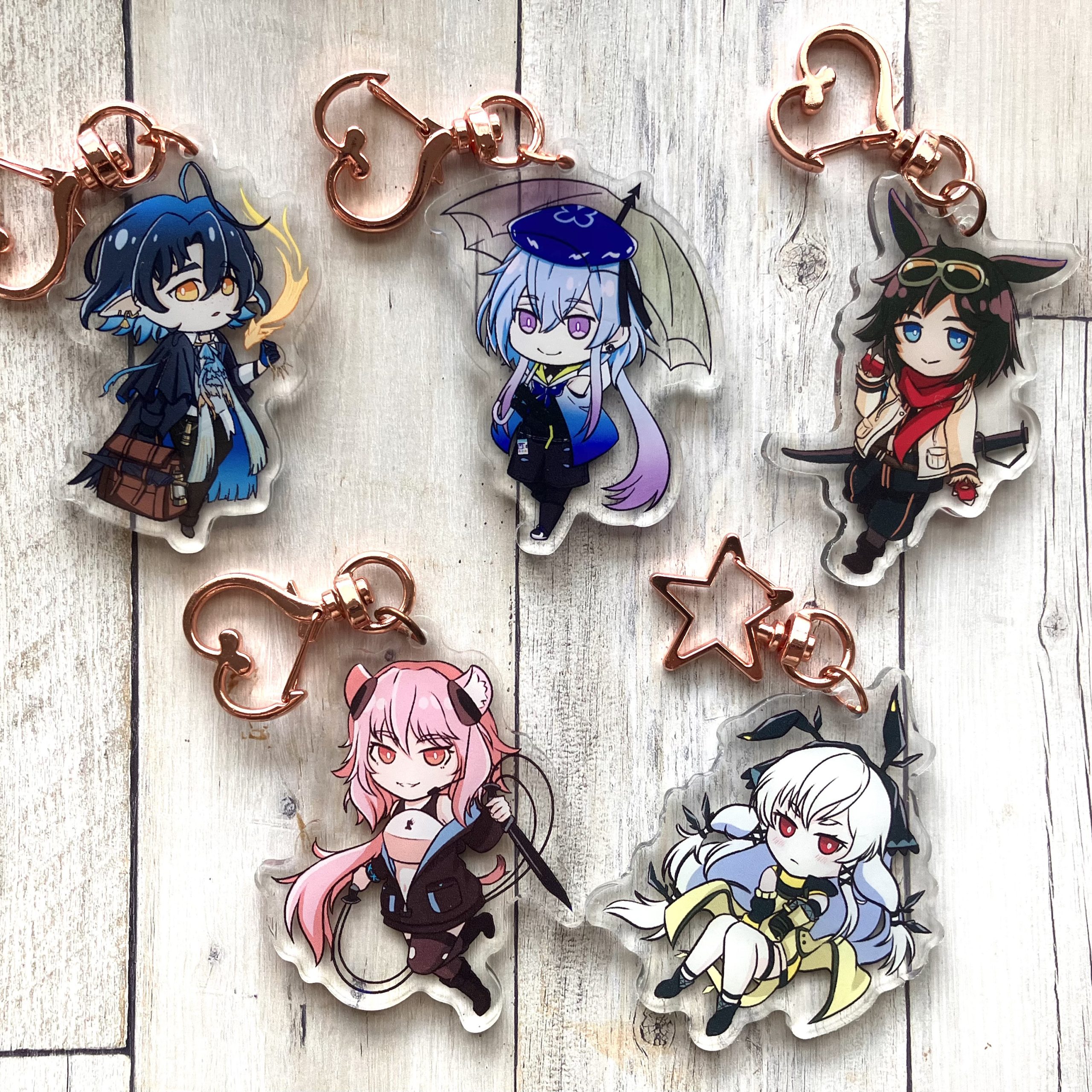 Five acrylic keychains of chibi-style anime characters with fantasy outfits and rose-gold clasps, arranged on a white wood background.