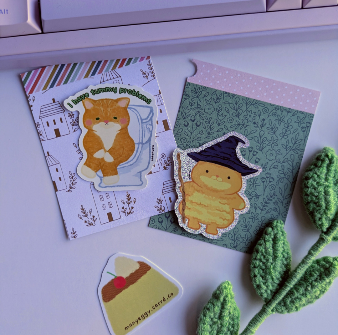 Cute cat and dessert stickers displayed on patterned paper near crocheted green leaves, including a cat with “I have tummy problems” text and a cat wearing a witch hat.